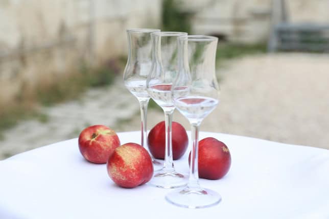 Three Palinka glasses standing next to fresh peaches on a white tablecloth