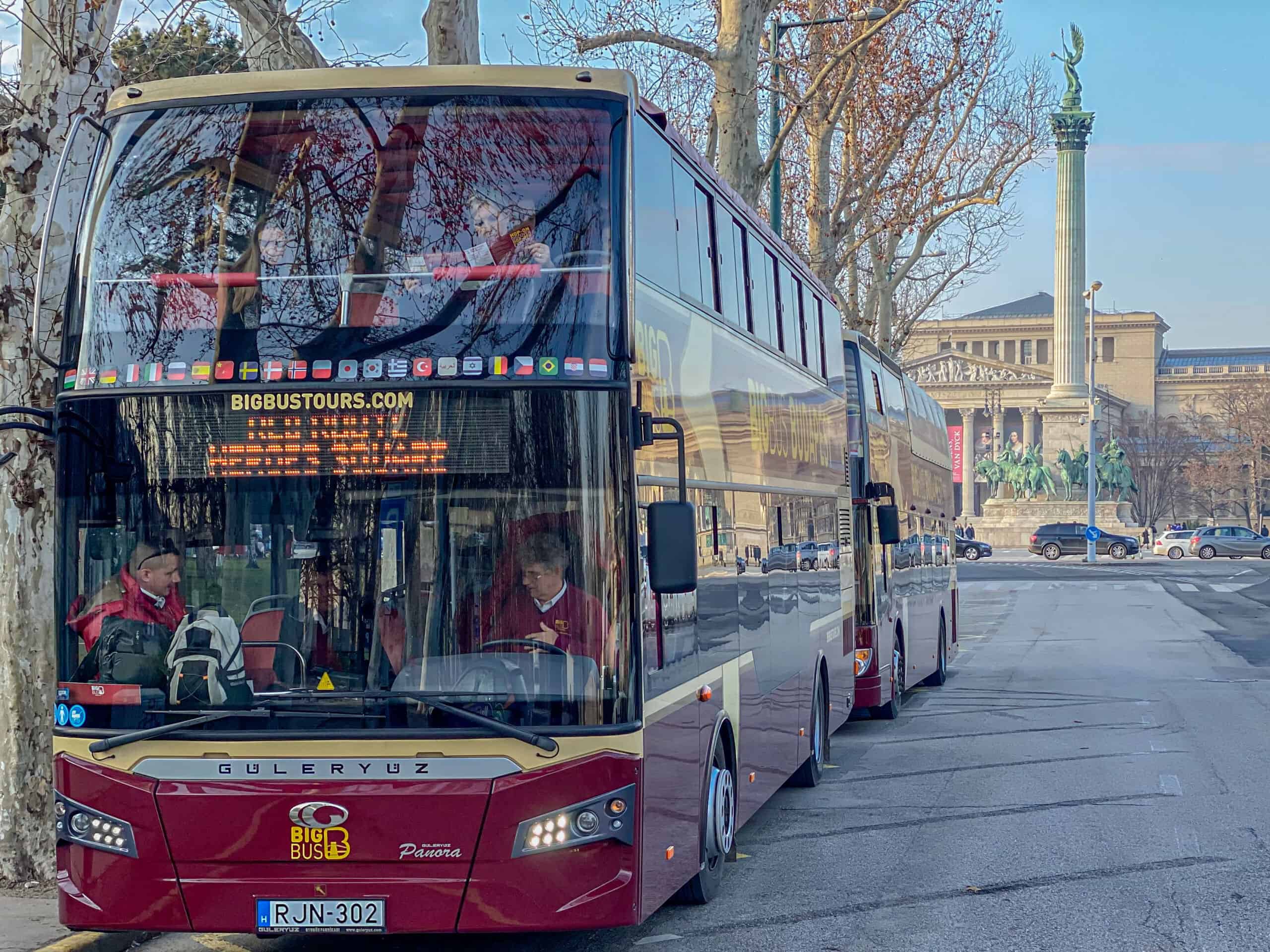 A red Big Bus sightseeing double-decker parked at Heroes' Square in Budapest with the Millennium Monument visible in the background.