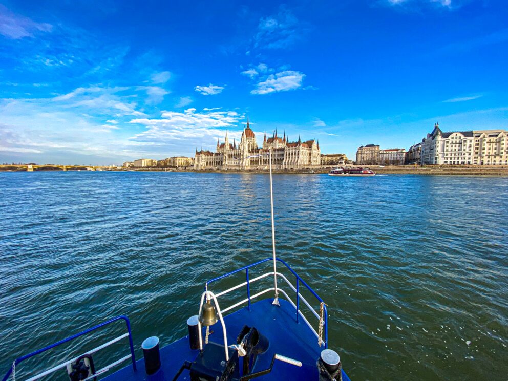 View of the Hungarian Parliament Building from the deck of a blue boat cruising on the Danube River in Budapest.