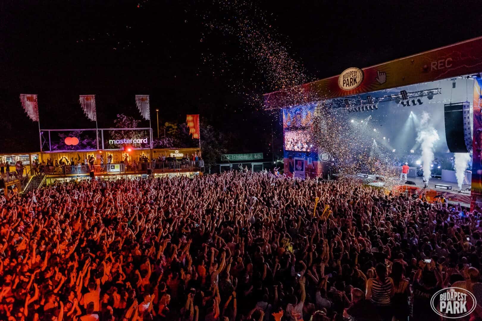 Thousands of people illuminated by pink and blue stage lights at an outdoor concert