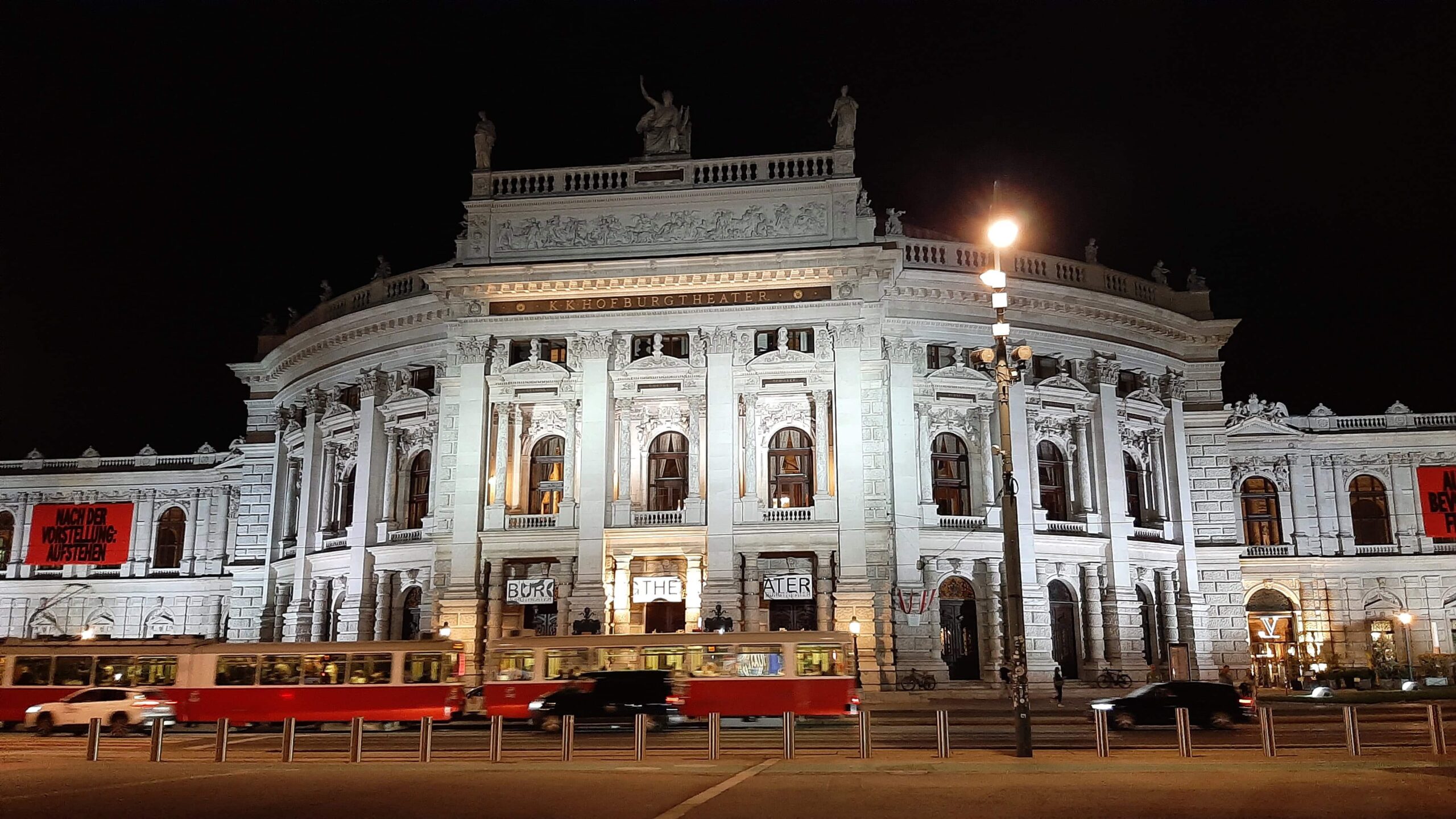 The illuminated Burgtheater in Vienna with a red tram passing by