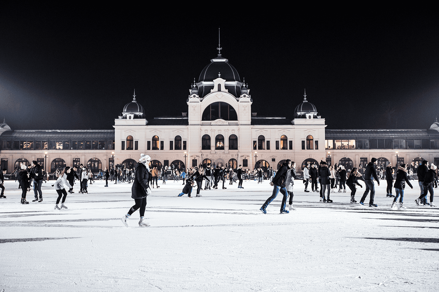 Large crowd of people ice skating at night at the illuminated City Park Ice Rink near Vajdahunyad Castle.