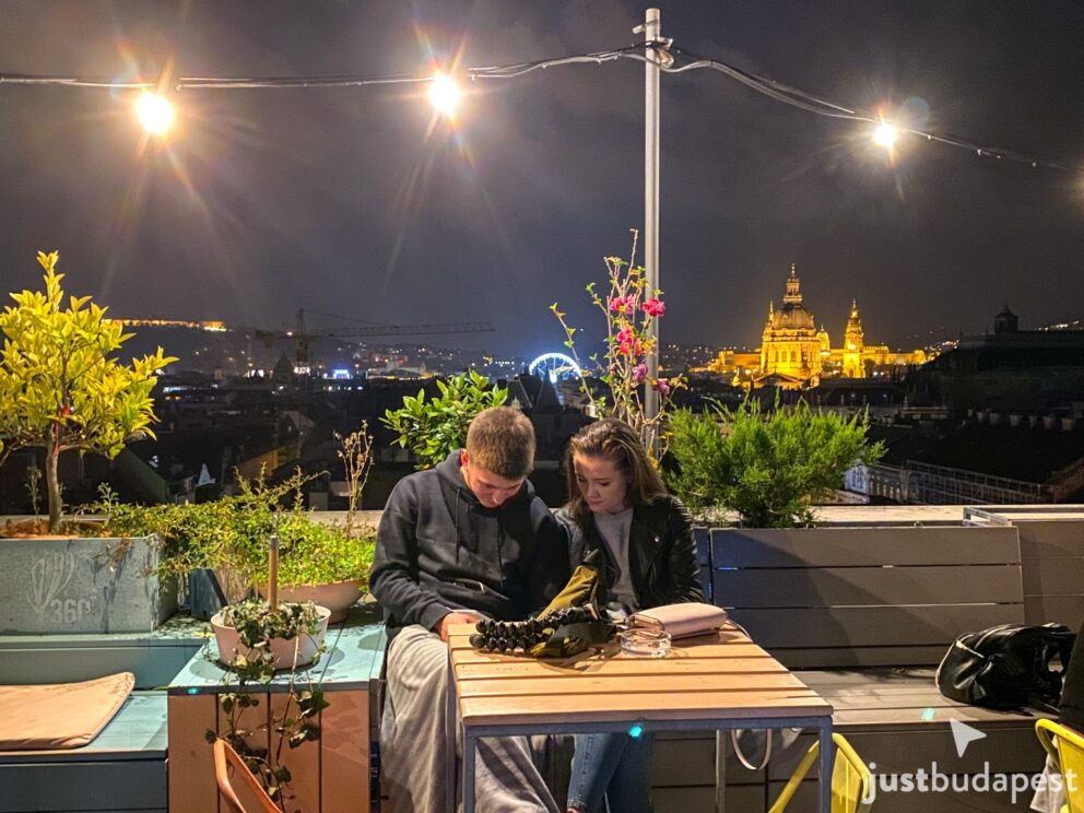 Couple enjoying a date at an outdoor rooftop table with illuminated Budapest skyline behind them