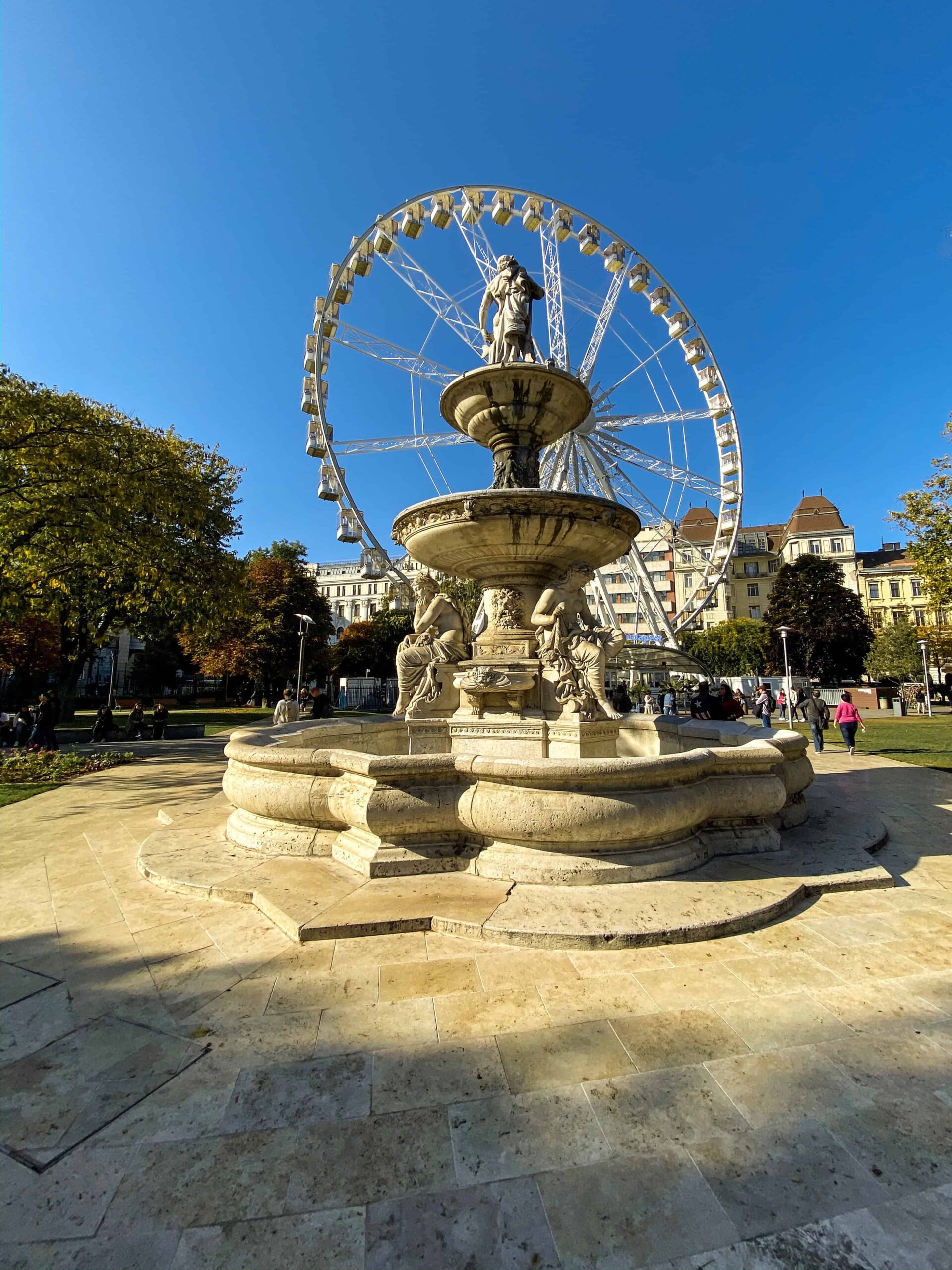 The historic Danubius Fountain spraying water with the modern Ferris wheel rising behind it
