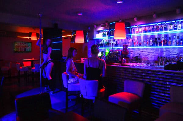 A dimly lit interior bathed in blue and red neon light showing a performer on a dancing pole behind a glowing bar