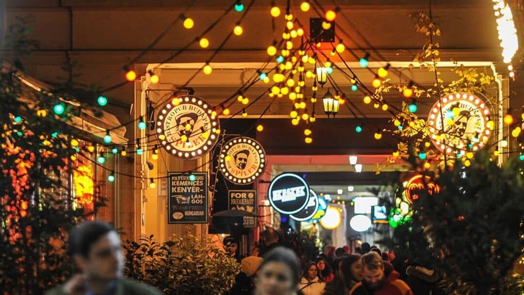 A highly crowded bustling nighttime street scene inside Gozsdu Udvar illuminated by dangling string lights
