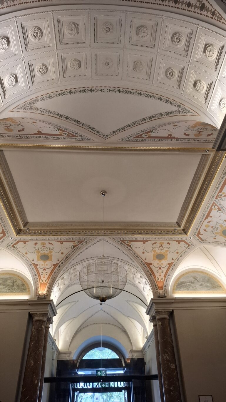An ornate painted vaulted ceiling decorated with delicate gold and beige patterns next to a modern chandelier