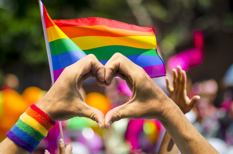 Two hands forming a heart shape in front of a blurred rainbow flag