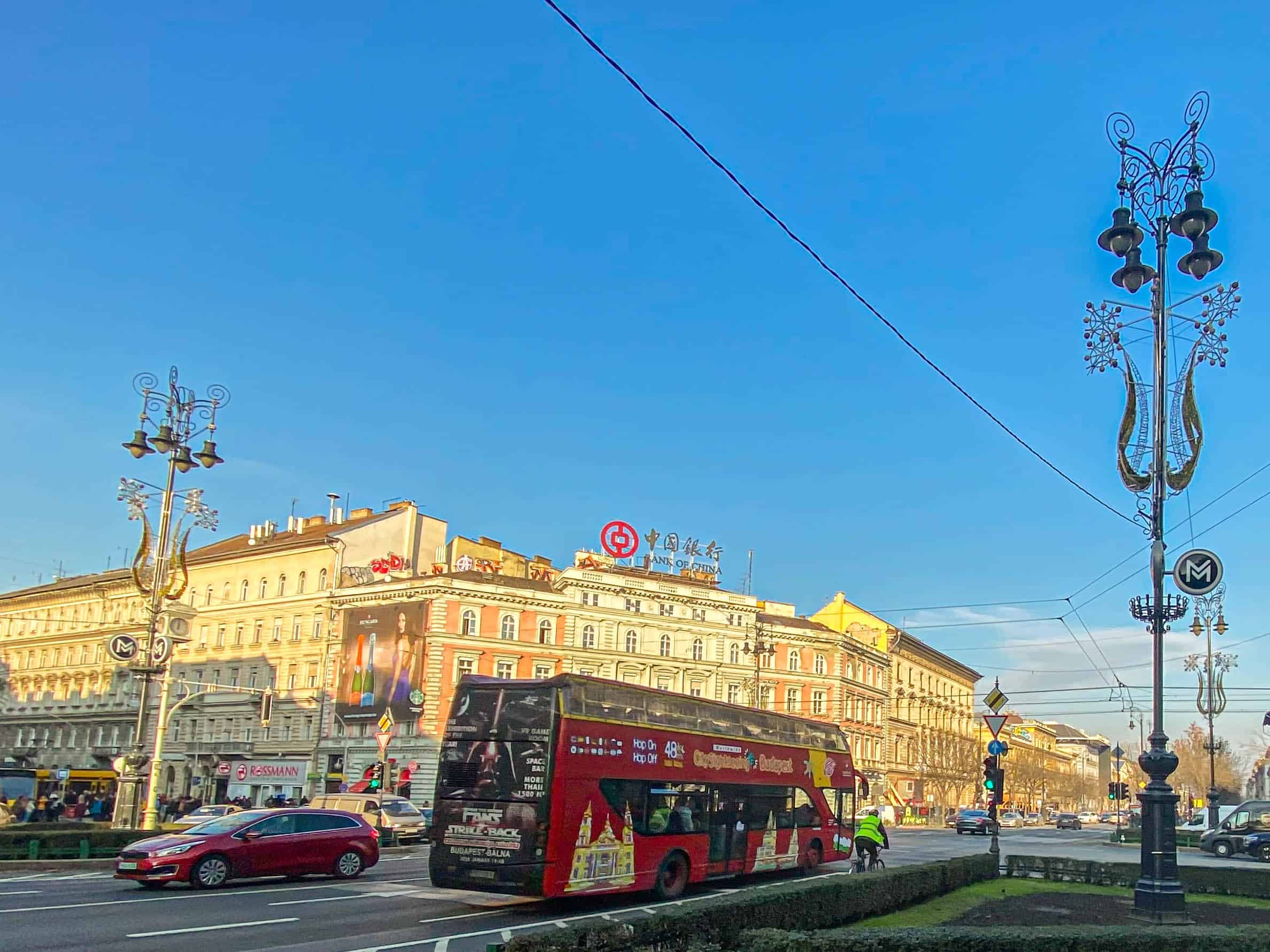 A red sightseeing bus driving through the busy Oktogon square on Andrássy Avenue