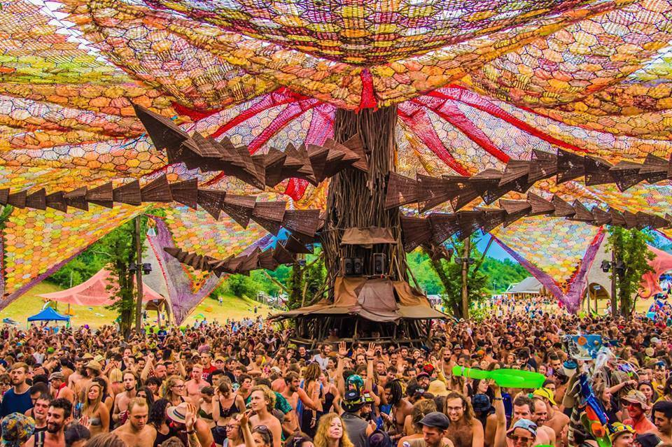 A massive crowd of festival-goers dancing under a giant colorful woven canopy at the main stage