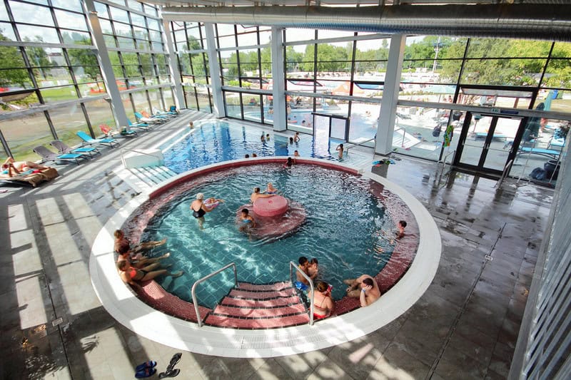 The modern interior of the Paskal Thermal Bath in Budapest with large glass windows and circular thermal pools.
