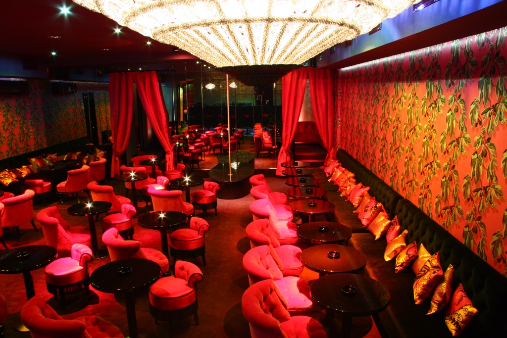 A brightly lit red club featuring a massive central sparkling chandelier and green leaf patterned wallpaper