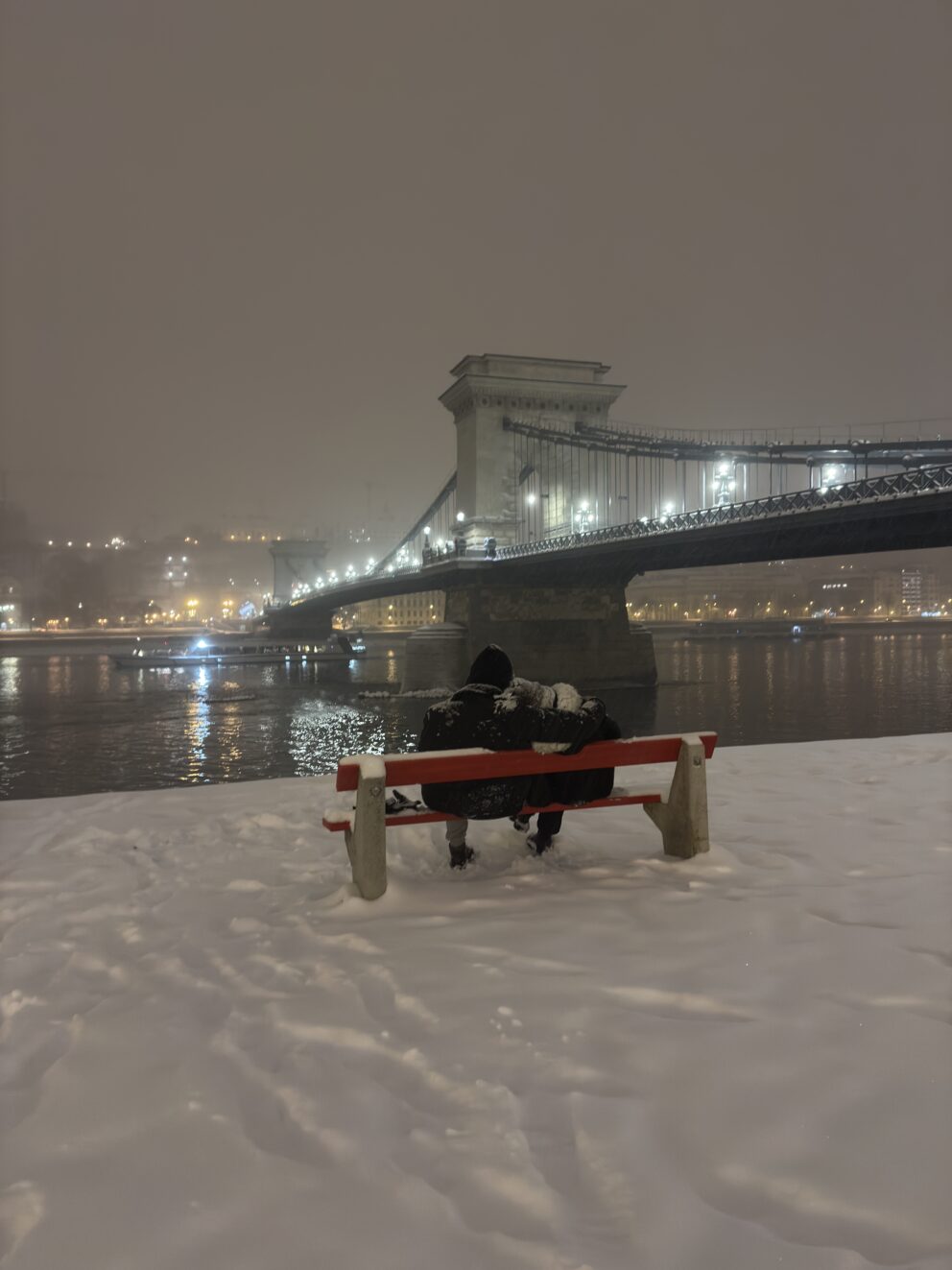 A couple sitting on a snowy bench enjoying the night view of the Chain Bridge