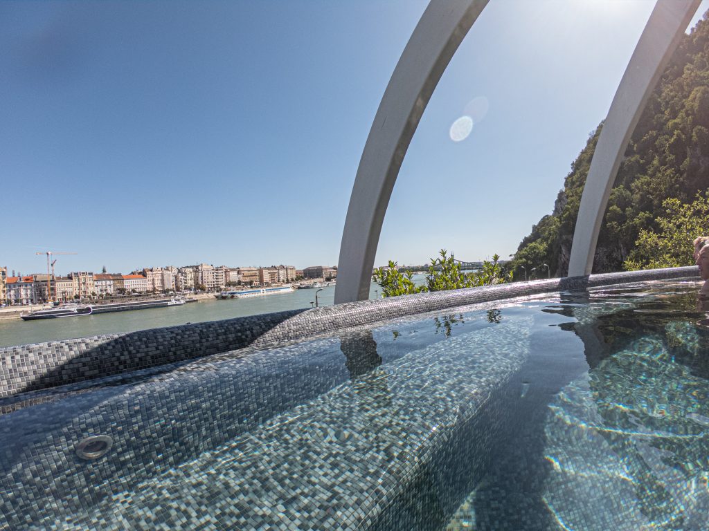 A high-angle view of the modern rooftop pool at Rudas Bath in Budapest, with the Danube River and city bridges in the background.
