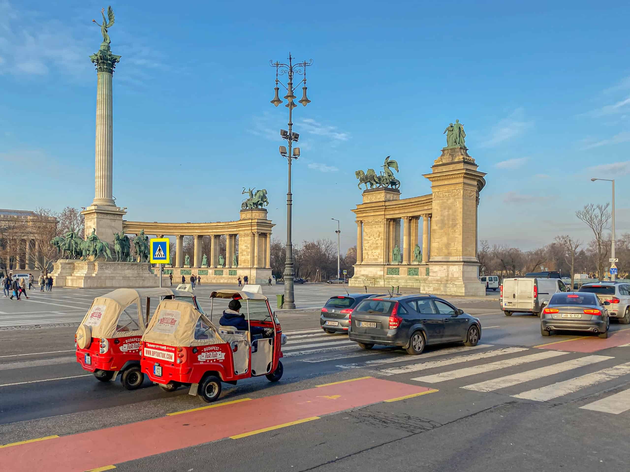 Tourists driving past the massive statues of Heroes' Square in a red Tuk-Tuk