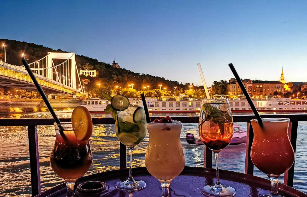 Enjoying colorful cocktails on the deck of a boat during a sunset cruise facing Buda Castle
