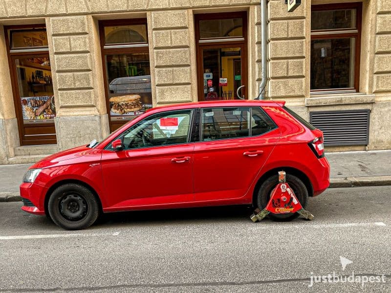 A red car immobilized with a heavy steel wheel clamp on a city street